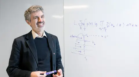 Photo of Yoshua Bengio standing next to a whiteboard with algorithms written on it 