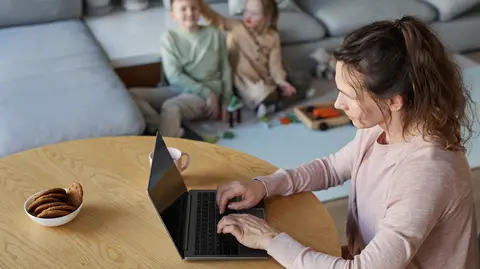 A woman working on a laptop at a table while two children play in the background.