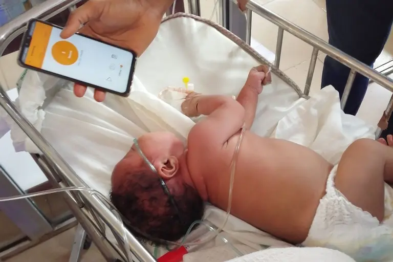 Un bébé dans un berceau à l'hôpital, avec quelqu’un qui monitorise ses pleurs à l’aide d’une application.
