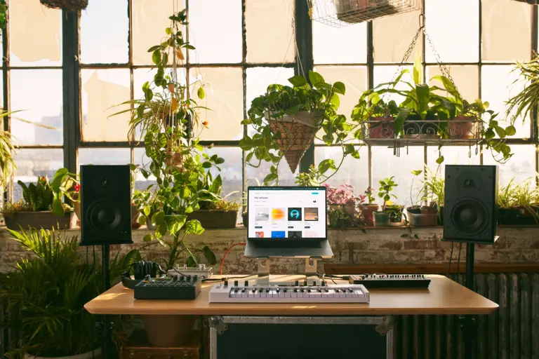 A music production setup on a wooden desk featuring a laptop, MIDI keyboard, and studio monitors, positioned in front of a large window filled with lush green houseplants.