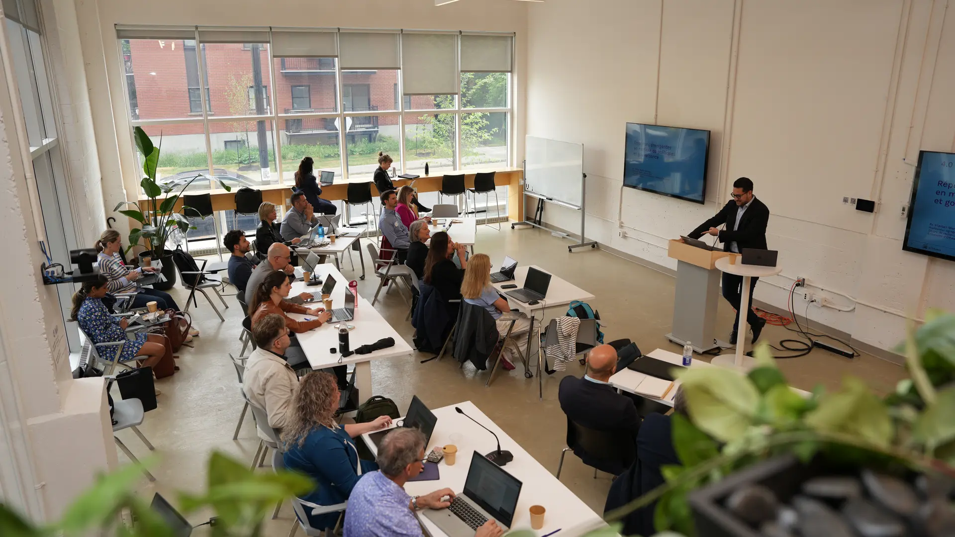 Picture of a group during a workshop where a speaker presents to a group of attendees in a bright, modern conference room with large windows and laptops on the tables.kshop