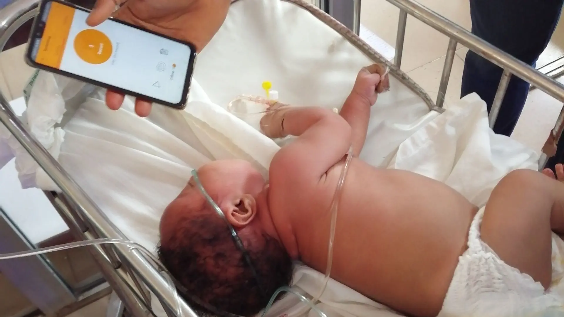 Un bébé dans un berceau à l'hôpital, avec quelqu’un qui monitorise ses pleurs à l’aide d’une application.