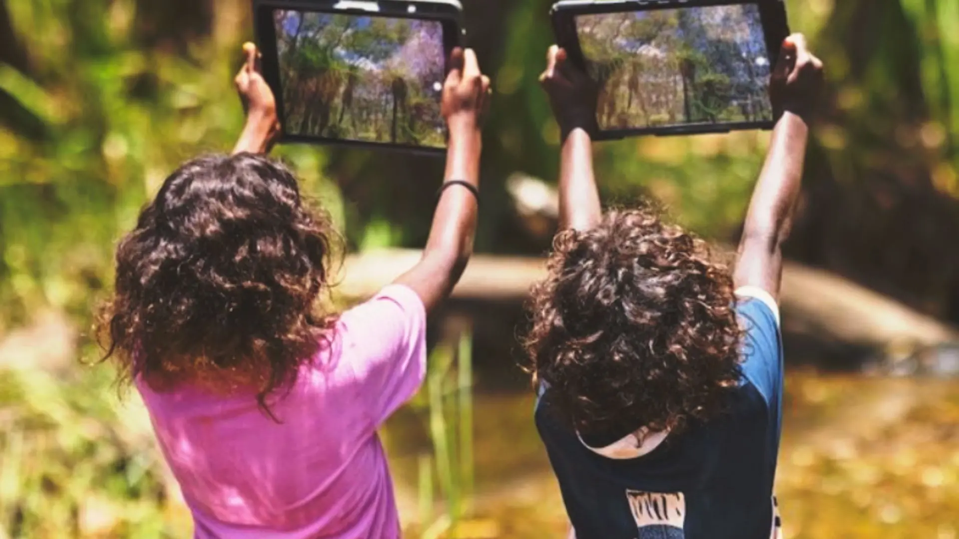 une photo de 2 enfants de dos à l'extérieur dans une forêt qui jouent à un jeu éducatif sur un ipad