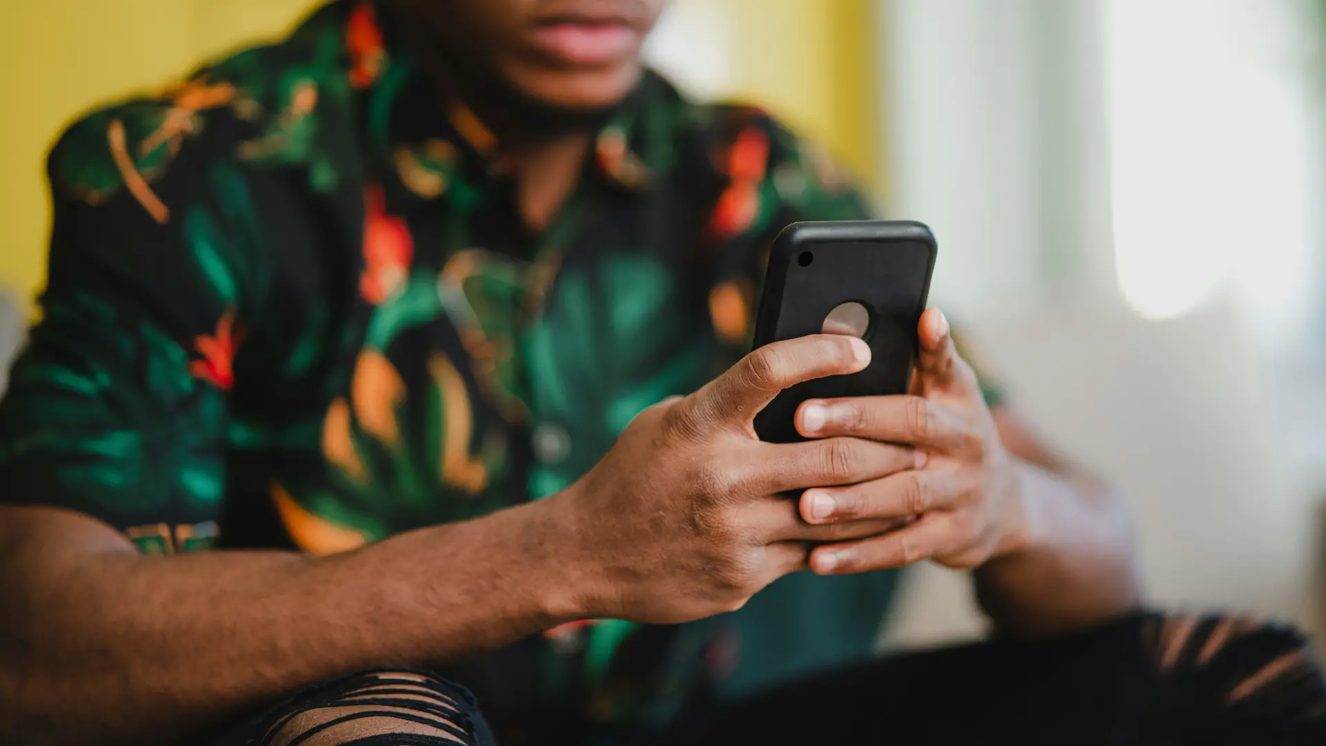 Photo d'un jeune homme assis regardant son cellulaire. Le fond est flou et le focus est mis sur ses mains qui tiennent son téléphone