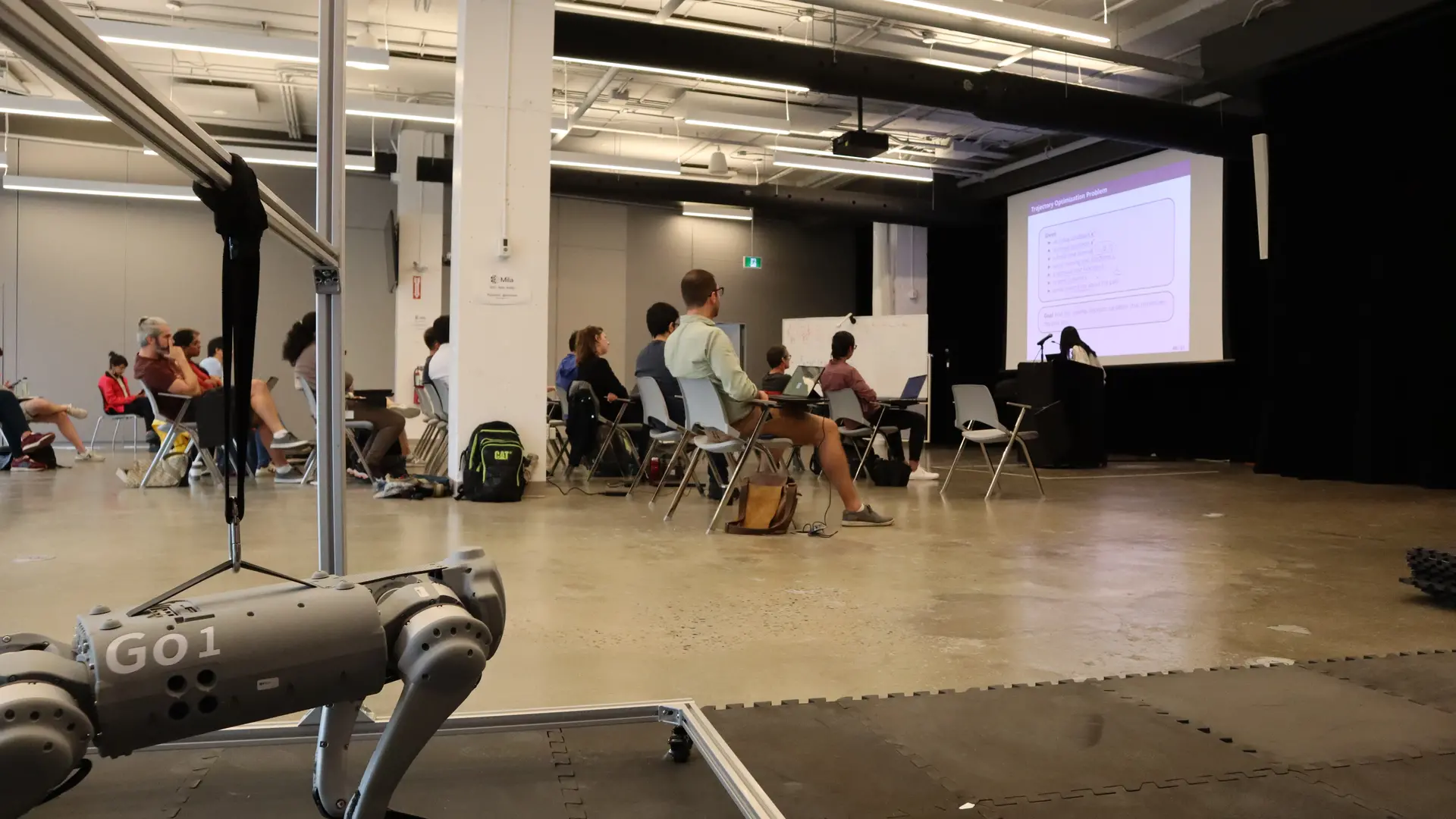 Students listen to a presentation, with a robot dog in the foreground. 