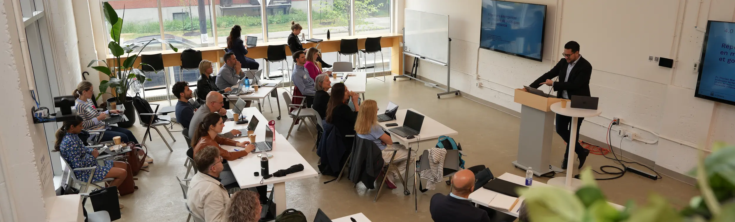 Un conférencier s'adresse à un groupe de participants dans une salle de conférence lumineuse et moderne, dotée de grandes fenêtres et de ordinateurs portables posés sur les tables