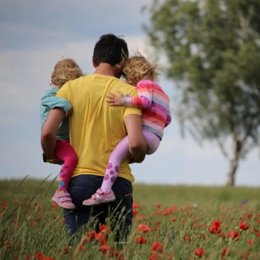 Man holding his two daughters in a field of red-petaled flowers.