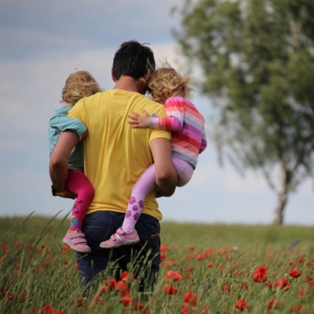 Homme tenant ses deux filles dans un champ de fleurs à pétales rouges.