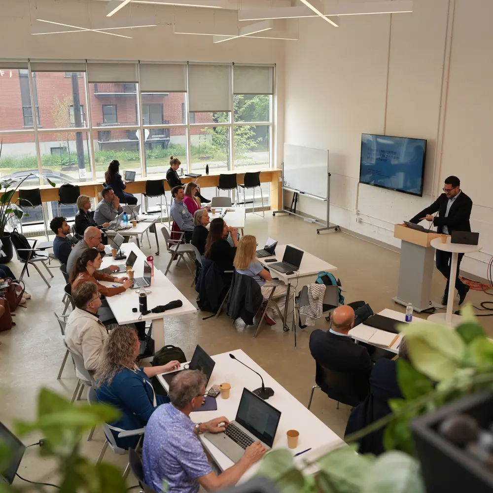 Participants of a worA speaker presents to a group of attendees in a bright, modern conference room with large windows and laptops on the tables.kshop